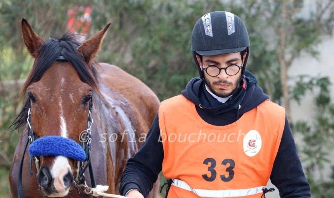 Amine Regaieg tient en main un cheval bai en compétition, dossard orange numéro 33 portant l’écusson de la fédération tunisienne, le cheval équipé d’une protection de chanfrein bleue.