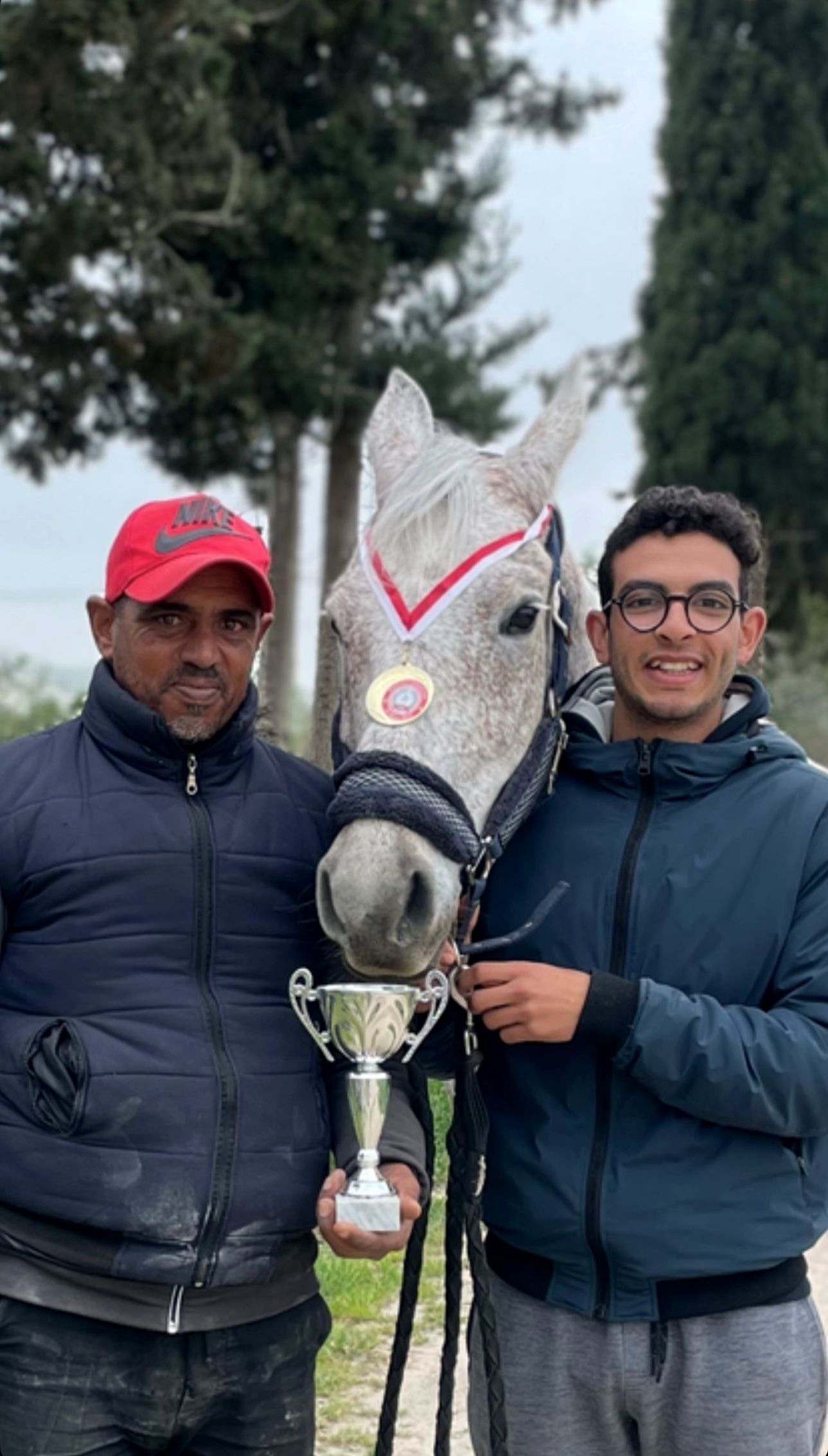Amine Regaieg et un coéquipier de part et d’autre d’un cheval gris portant une médaille sur ruban rouge et blanc, une coupe tenue entre eux.